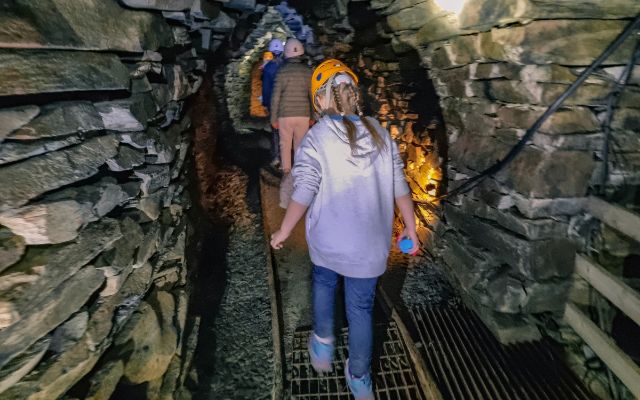 people in hard hats walking along tracks to torchlight inside a slate mine tunnel