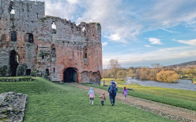 : grey and red stone castle ruins sit next to a river with a family walking towards them across grass