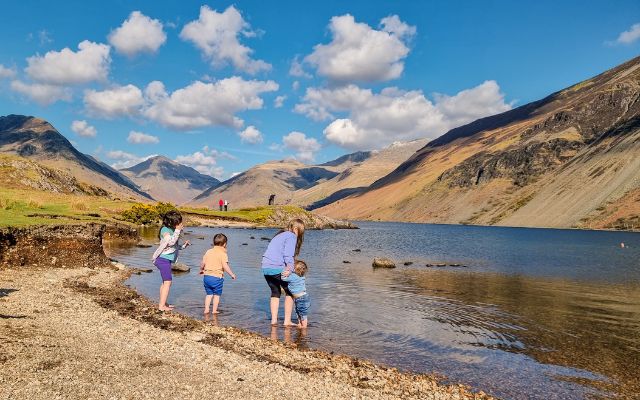 four children are paddling at the water’s edge of Wast Water with the Lakeland fells beyond and blue sky above