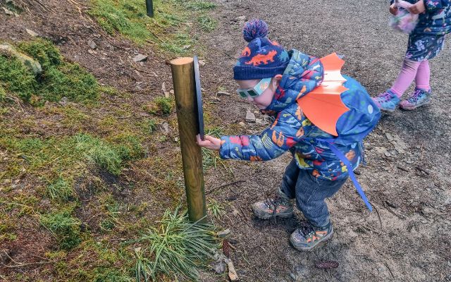 a boy in a dinosaur coat wearing orange dragon wings and 3D glasses is looking at a sign on the Zog Trail at Whinlatter Forest 