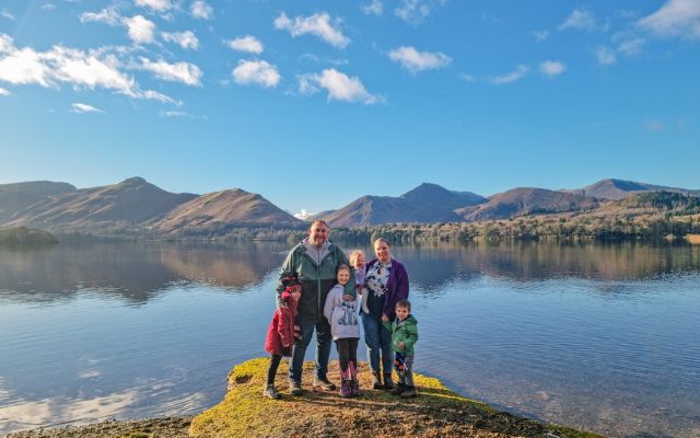 a family of 6 is standing smiling st the camera with Derwentwater and the Lakeland fells behind them beneath a blue sky